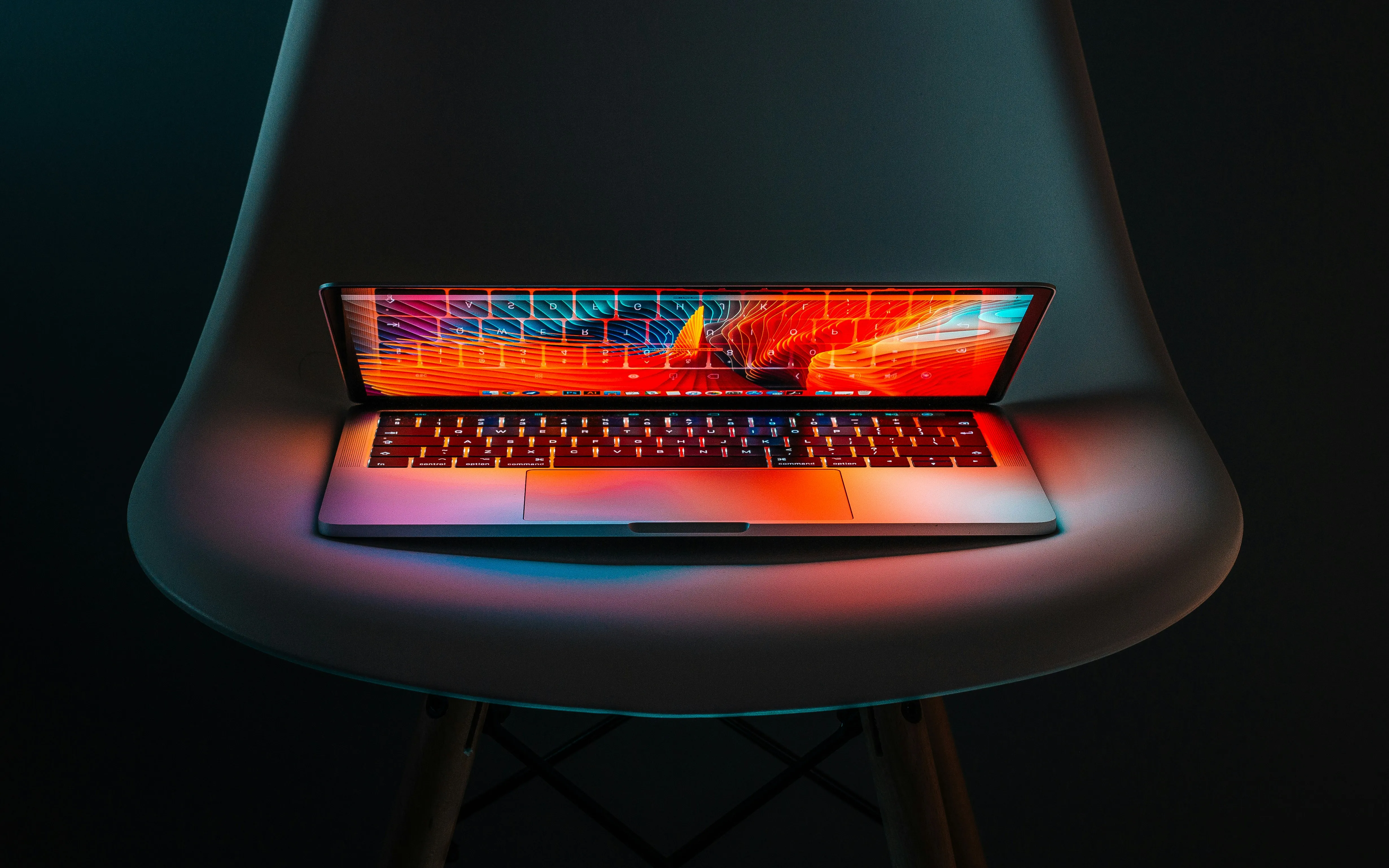 A macbook sitting on a chair, half closed with colors pouring out from the display on a badk background.